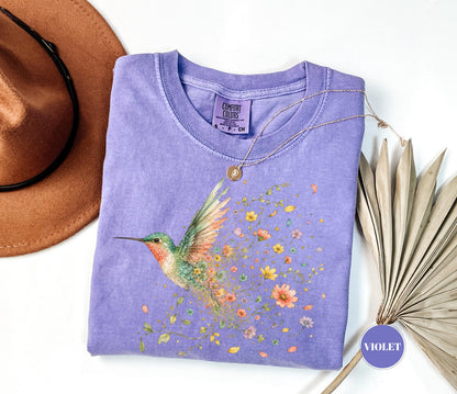 May include: A violet short-sleeve t-shirt with a Comfort Colors label, featuring a graphic of a hummingbird with colorful wings and a trail of small flowers. A thin gold necklace with a small circular pendant rests on the neckline of the shirt.