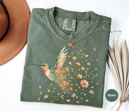 May include: A moss green crew neck t-shirt featuring a graphic of a hummingbird with wings transitioning into a trail of colorful flowers and leaves. The inner neck label reads Comfort Colors, 100% cotton, made in Honduras, size S-CH.