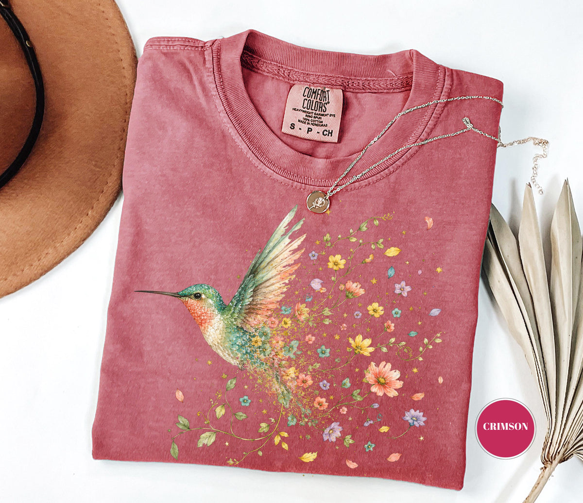 May include: A crimson short-sleeve t-shirt featuring a graphic of a hummingbird with wings transitioning into a trail of colorful flowers. The shirt has a crew neckline and a Comfort Colors brand label at the inner neck. A thin gold chain necklace is present.