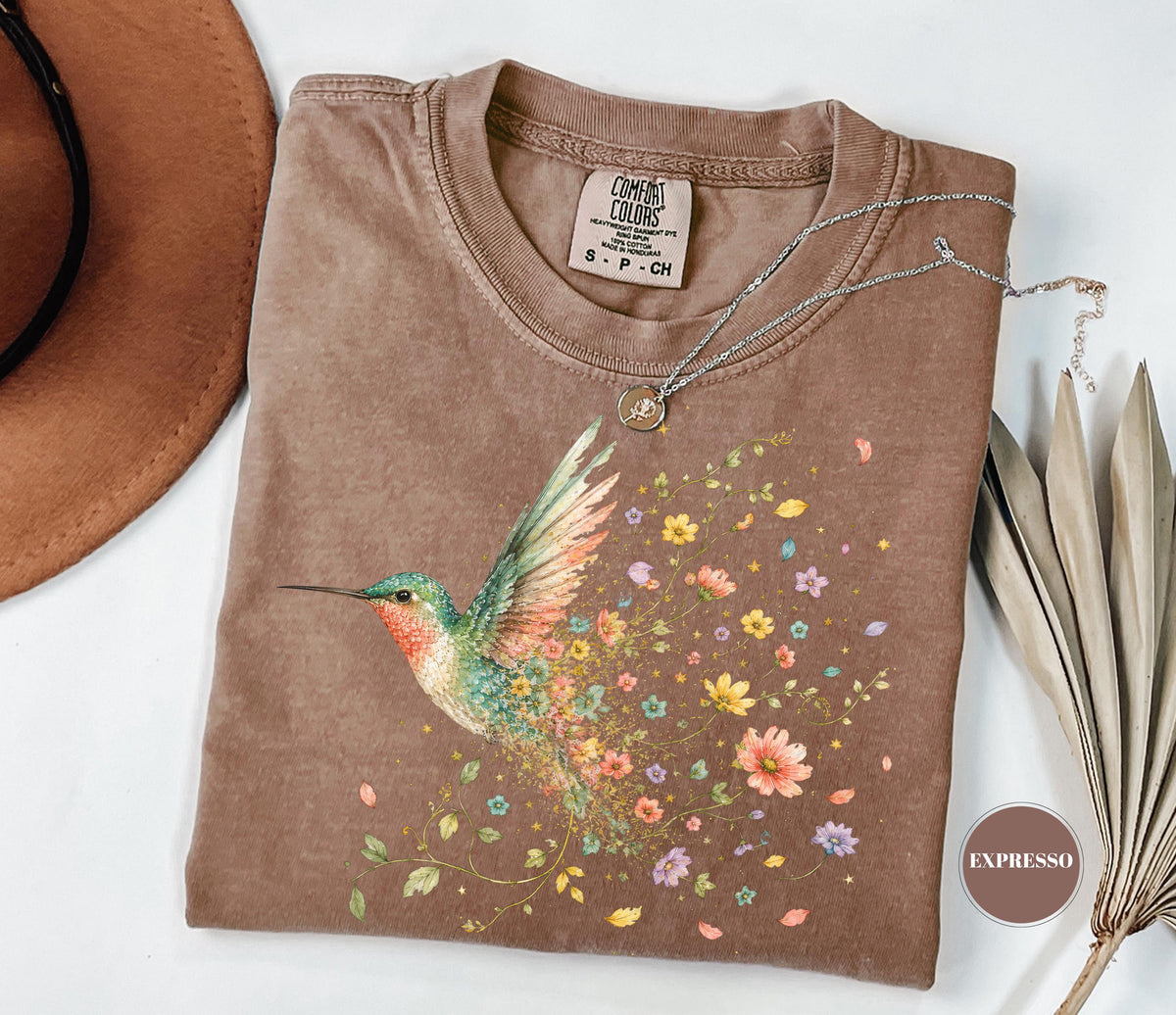May include: A brown short-sleeve t-shirt featuring a graphic of a hummingbird with green, orange, and yellow feathers. The bird is surrounded by a trail of colorful flowers and leaves. The shirt has a crew neckline and a Comfort Colors brand label.