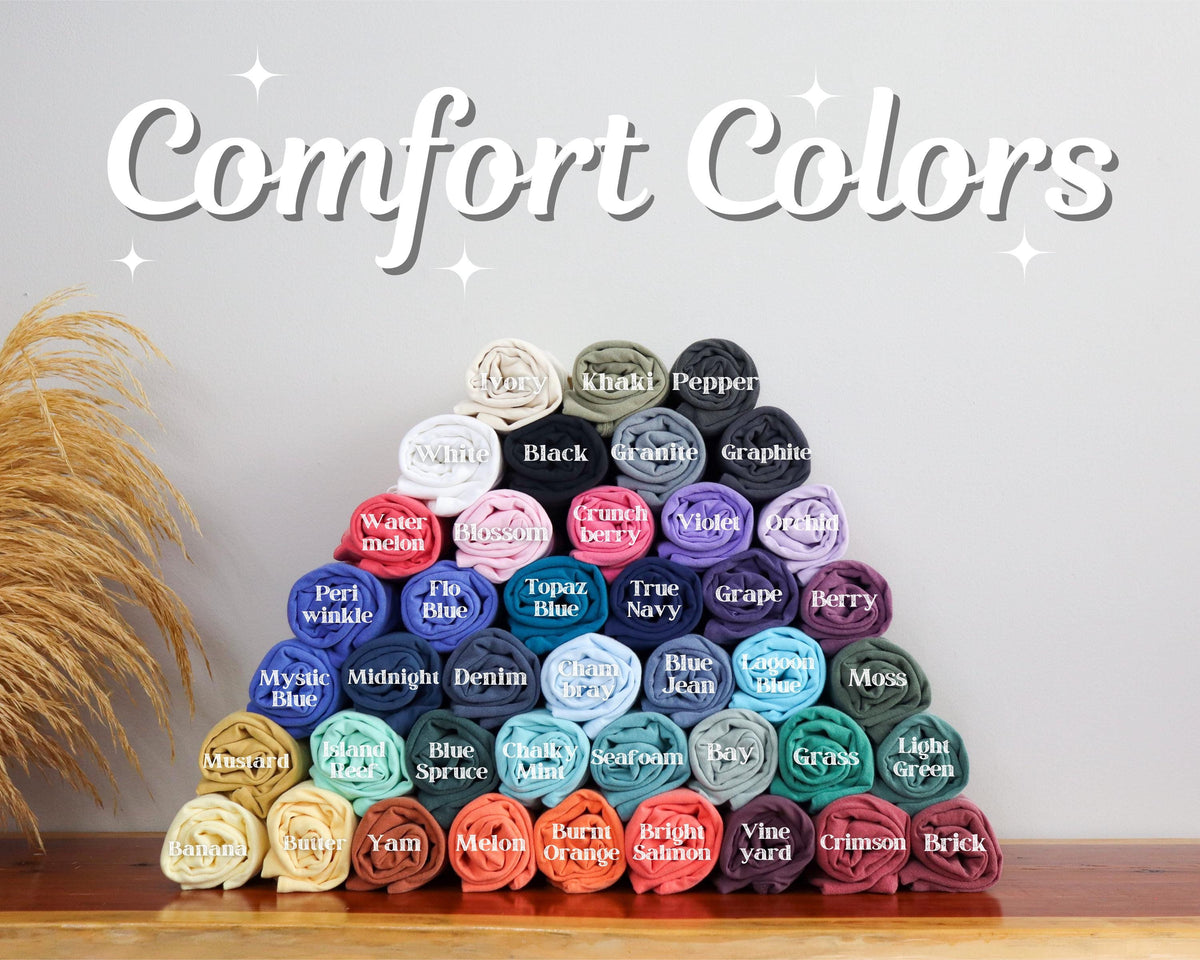 May include: A pyramid of rolled t-shirts in various colors labeled with text including Ivory, Khaki, Pepper, Black, Granite, Graphite, Watermelon, Blossom, Crunchberry, Violet, Orchid, Periwinkle, Flo Blue, Topaz Blue, True Navy, Grape, Berry, and more.