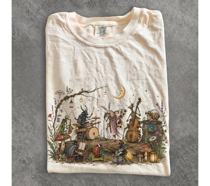 May include: A cream-colored t-shirt featuring a detailed graphic print of various insects playing musical instruments on a log. The illustration includes a beetle on drums, a moth singing, a snail with headphones, and a ladybug playing a flute.
