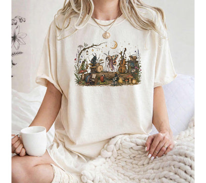 May include: An off-white short-sleeve t-shirt featuring a detailed, colorful graphic print of various insects playing musical instruments in a forest setting, including a beetle on drums, a moth on a cello, and a snail, surrounded by mushrooms.