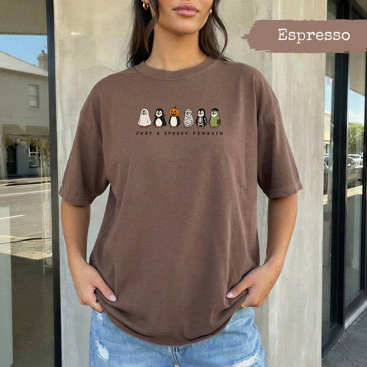 Brown t-shirt with six cartoon penguins in Halloween costumes and text "JUST A SPOOKY PENGUIN".