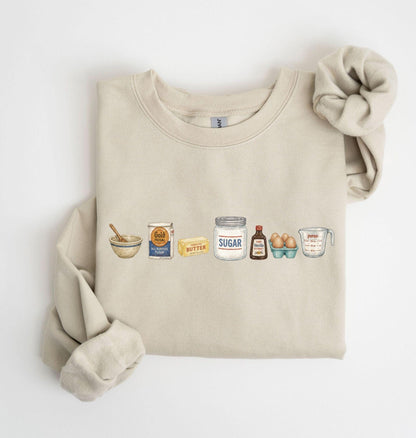 May include: A beige crewneck sweatshirt featuring a graphic print of baking ingredients in a row, including a mixing bowl with a spoon, a bag of Gold Medal flour, a stick of butter, a jar labeled SUGAR, vanilla extract, eggs, and a Pyrex measuring cup.