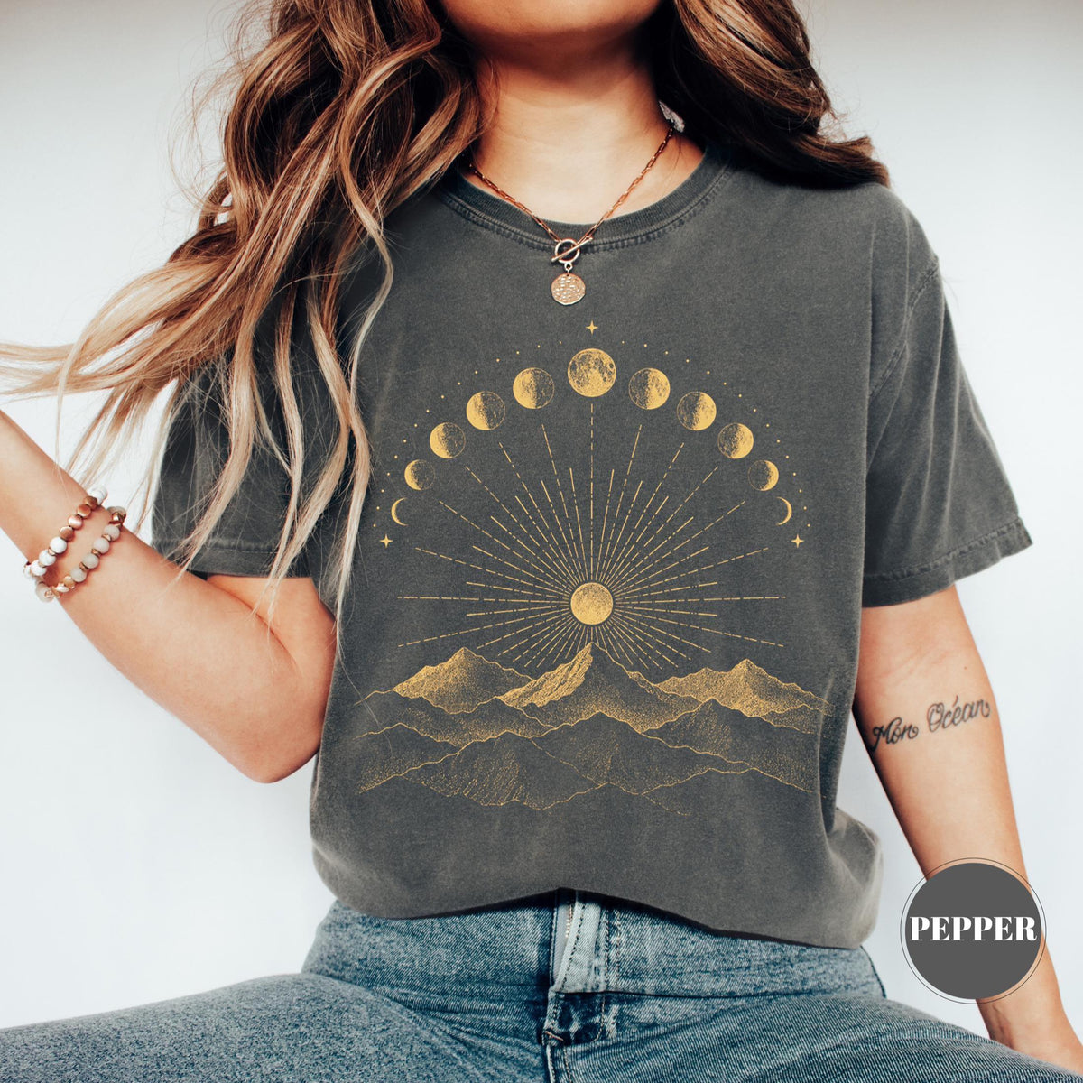 May include: A charcoal gray t-shirt featuring a gold-toned graphic print of a mountain range beneath a sunburst and a crescent-to-full moon phase cycle. The design includes fine lines radiating from the sun, creating a celestial landscape.