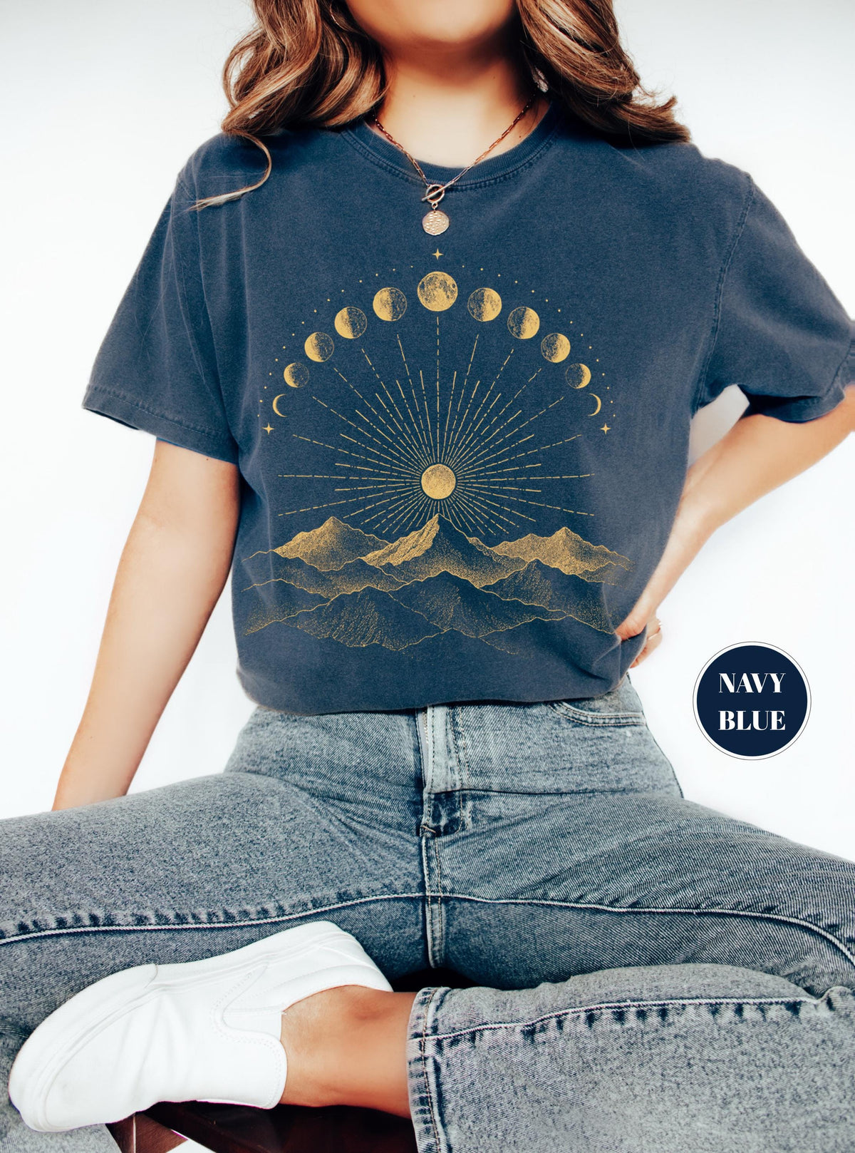 May include: A navy blue t-shirt featuring a gold graphic print of a mountain range beneath a sun with radiating lines and a semi-circular arrangement of moon phases. The shirt has short sleeves and a crew neckline, with the text NAVY BLUE in a circle.
