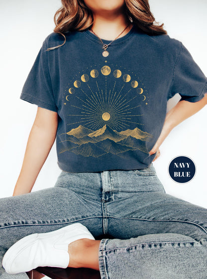 May include: A navy blue t-shirt featuring a gold graphic print of a mountain range beneath a sun with radiating lines and a semi-circular arrangement of moon phases. The shirt has short sleeves and a crew neckline, with the text NAVY BLUE in a circle.
