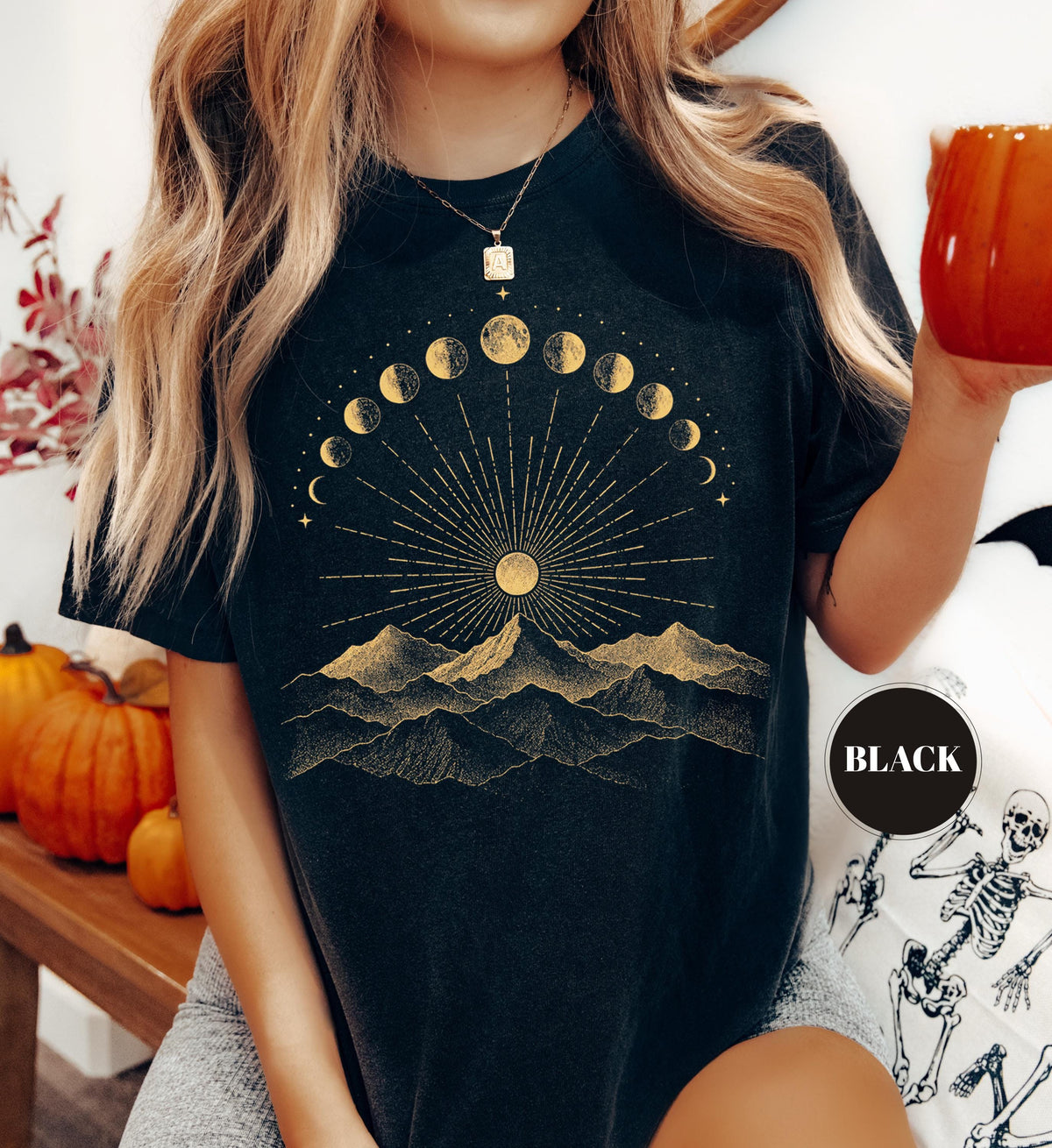 May include: A black t-shirt featuring a gold graphic print of a mountain range beneath a sun with radiating light rays and a crescent-to-full moon phase cycle arching across the top. A circular badge on the lower right displays the word BLACK.