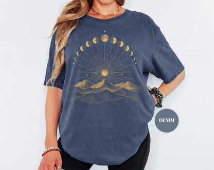 May include: A denim blue short-sleeve t-shirt featuring a gold-toned graphic print on the chest. The design displays a mountain range beneath a sunburst and an arc of moon phases. A circular badge on the right side contains the text DENIM.