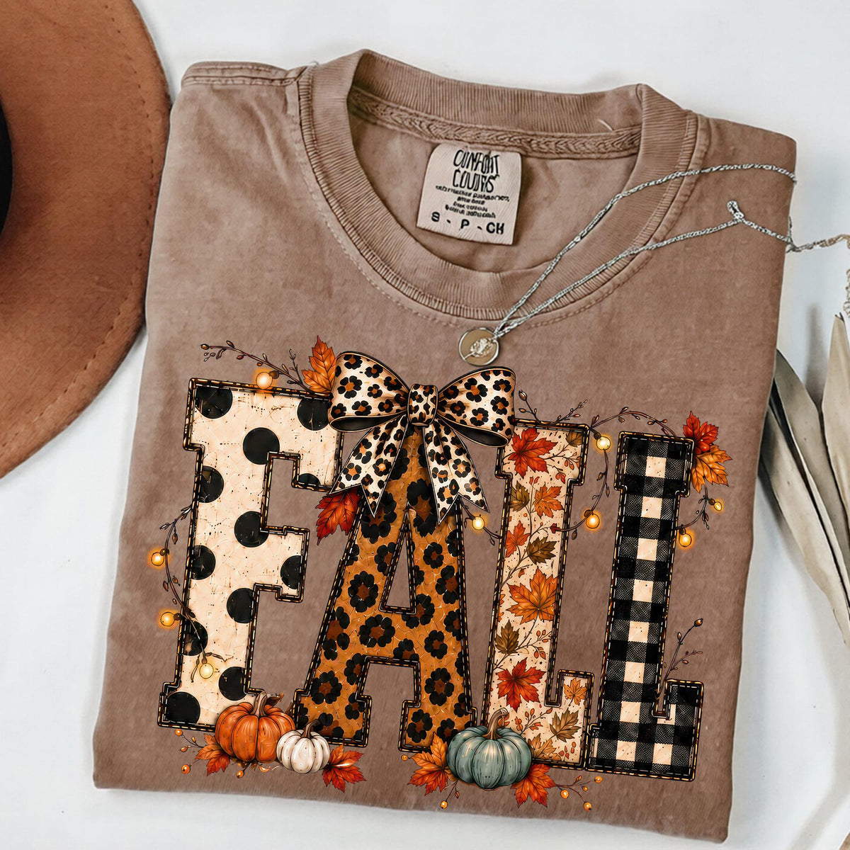 May include: A tan short-sleeved t-shirt featuring the word FALL in large block letters. Each letter has a different pattern: polka dots, leopard print, autumn leaves, and black plaid. A leopard print bow sits above the letters with pumpkins.