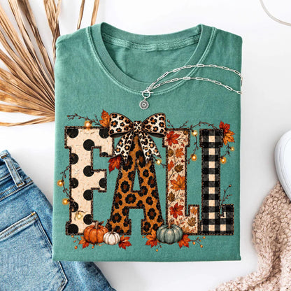 May include: A sage green t-shirt featuring the word FALL in large block letters. Each letter has a different pattern: black polka dots, leopard print, autumn leaves, and black buffalo plaid. A leopard print bow sits atop the letter A.