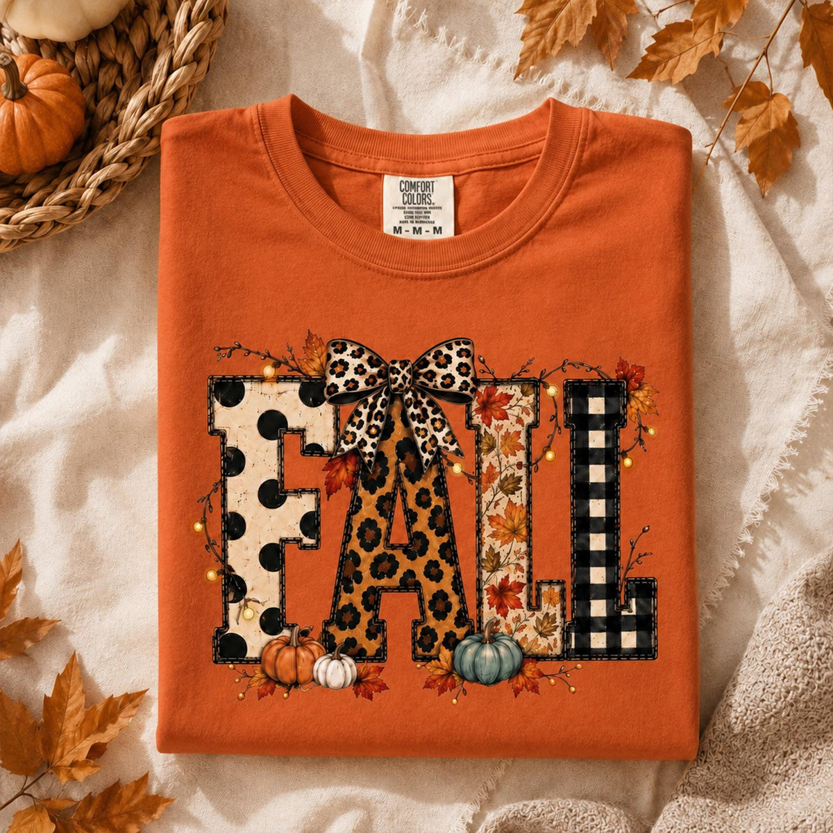 May include: An orange t-shirt featuring the word FALL in large block letters. Each letter has a different pattern: black polka dots, leopard print, autumn leaves, and black buffalo plaid. A leopard print bow sits atop the letter A.
