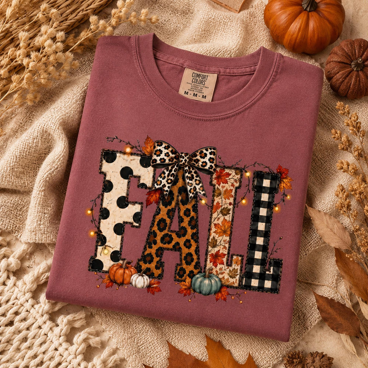 May include: A mauve crewneck t-shirt featuring the word FALL in large block letters. Each letter has a different pattern: black polka dots, leopard print, autumn leaves, and black buffalo plaid. A leopard print bow sits atop the letters.