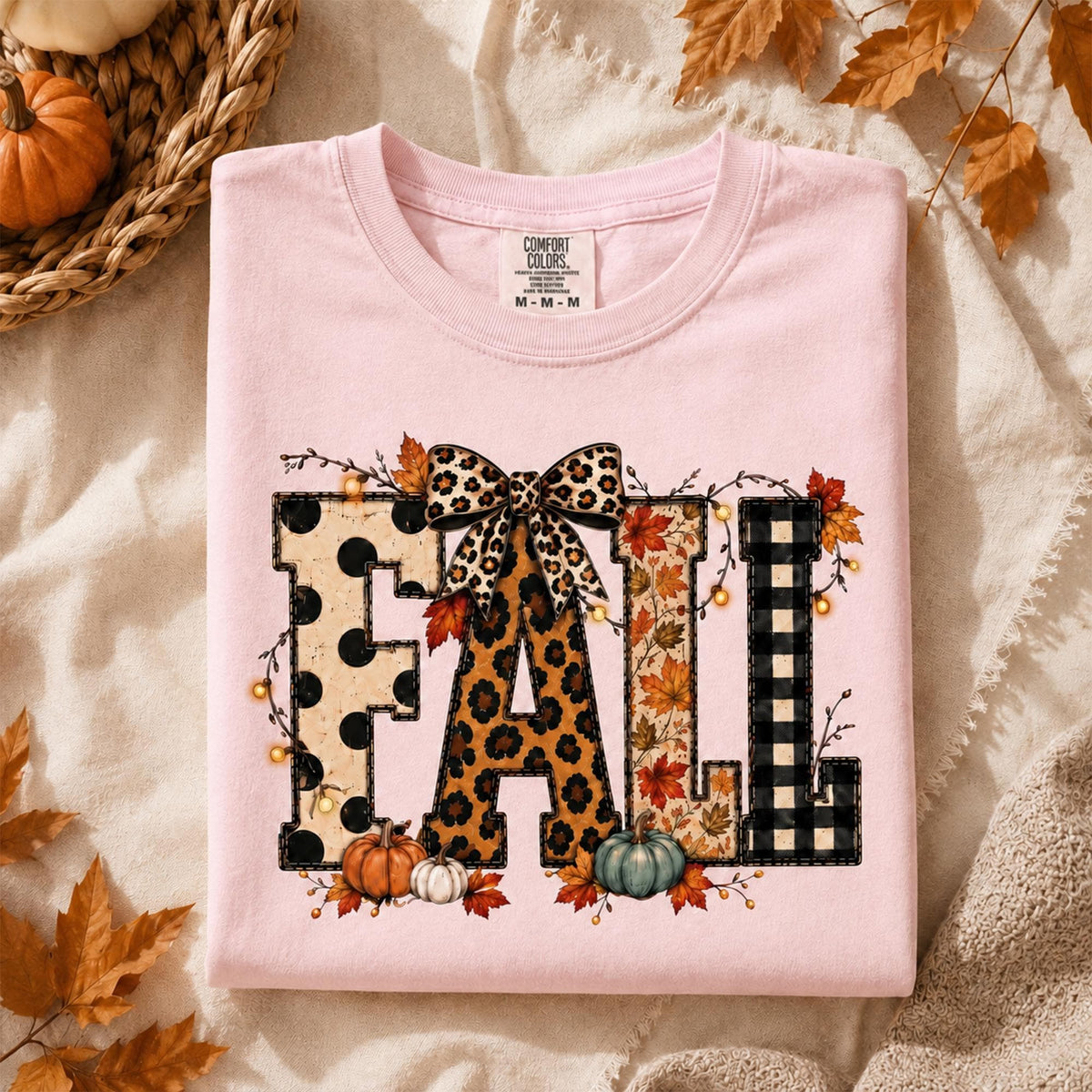 May include: A light pink t-shirt featuring the word FALL in large block letters. Each letter has a different pattern: black polka dots, leopard print, autumn leaves, and black buffalo plaid. A leopard print bow sits atop the letters.