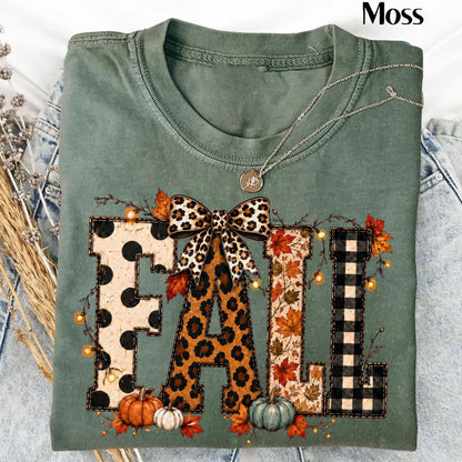 May include: A sage green t-shirt featuring the word FALL in large block letters. Each letter has a different pattern: black polka dots, leopard print, autumn leaves, and black buffalo plaid. A leopard print bow sits atop the letters.