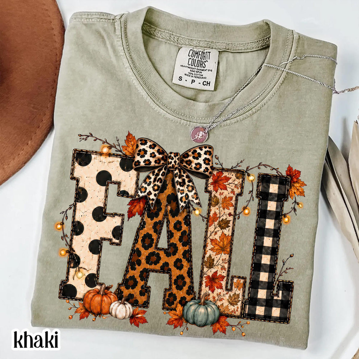 May include: A sage green short-sleeve t-shirt featuring the word FALL in large block letters. The letters have polka dot, leopard print, autumn leaf, and buffalo plaid patterns. A leopard print bow sits above the A, with pumpkins below.