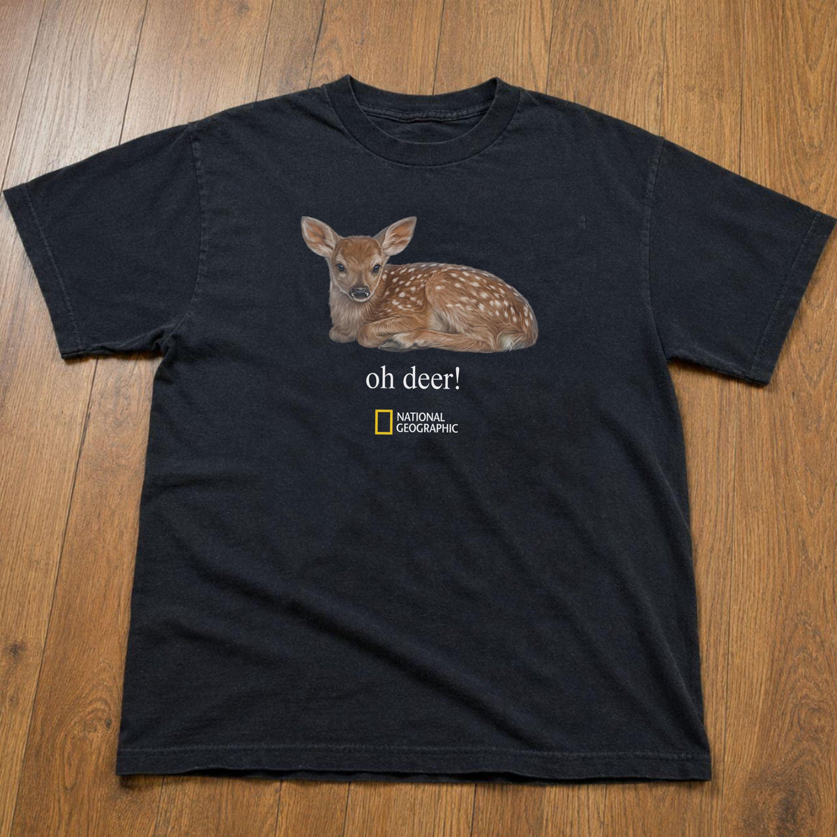 May include: A black short-sleeved t-shirt featuring a graphic of a brown spotted fawn lying down. Below the fawn, the text reads "oh deer!" in white, followed by the yellow and white National Geographic logo centered on the chest area.