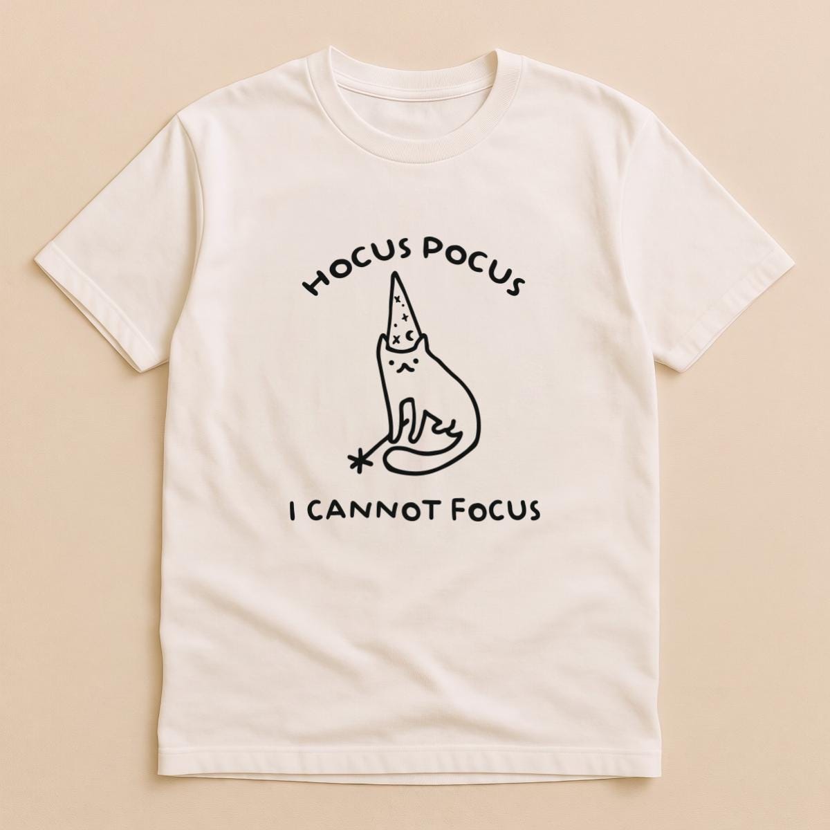 May include: A cream-colored short-sleeve t-shirt featuring a black line drawing of a cat wearing a wizard hat and holding a magic wand. The text HOCUS POCUS is printed above the cat, and the text I CANNOT FOCUS is printed below the cat.