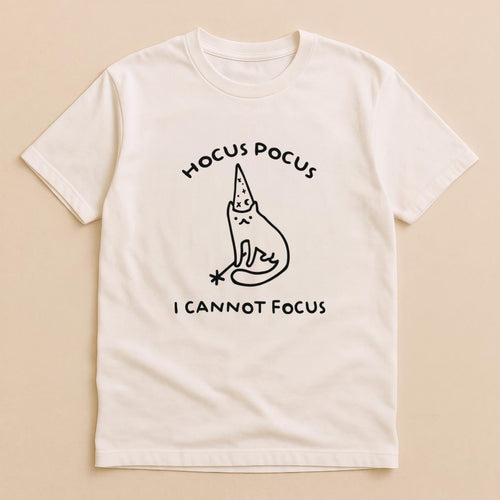 May include: A cream-colored short-sleeve t-shirt featuring a black line drawing of a cat wearing a wizard hat and holding a magic wand. The text HOCUS POCUS is printed above the cat, and the text I CANNOT FOCUS is printed below the cat.