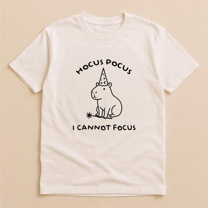 May include: A cream-colored short-sleeve t-shirt featuring a black line drawing of a capybara wearing a wizard hat and holding a magic wand. The text HOCUS POCUS is printed above the capybara, and I CANNOT FOCUS is printed below the graphic.