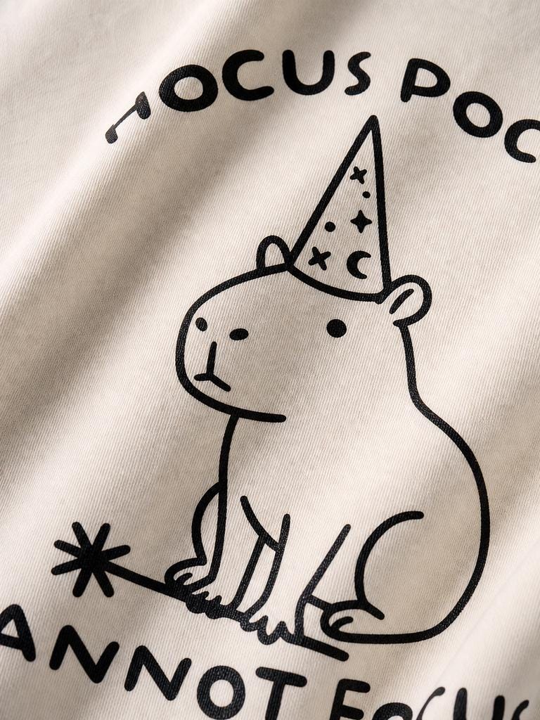 May include: A cream-colored fabric featuring a black line-art illustration of a capybara wearing a wizard hat decorated with stars and a crescent moon. The text HOCUS POCUS is printed above, and the text ANNOT FOCUS is printed below the capybara.