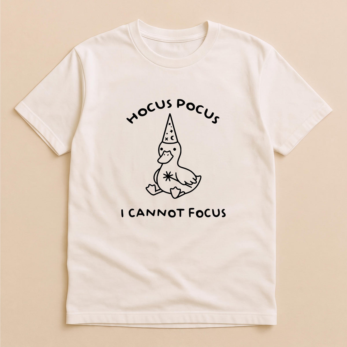 May include: A cream-colored short-sleeve t-shirt featuring a black line-art graphic of a duck wearing a wizard hat decorated with stars and moons. The text HOCUS POCUS is printed above the duck, and I CANNOT FOCUS is printed below the duck.