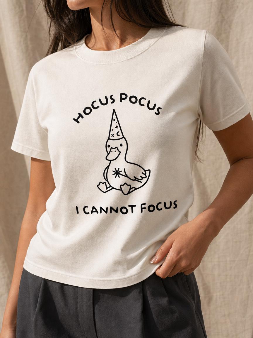 May include: A cream-colored short-sleeve t-shirt featuring a black line-art graphic of a duck wearing a wizard hat decorated with stars and moons. The text "HOCUS POCUS" is printed above the duck, and "I CANNOT FOCUS" is printed below the duck.