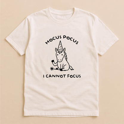 May include: A cream-colored short-sleeve t-shirt featuring a black line-art graphic of a unicorn wearing a wizard hat and holding a magic wand. The text HOCUS POCUS is printed above the unicorn, and the text I CANNOT FOCUS is printed below.