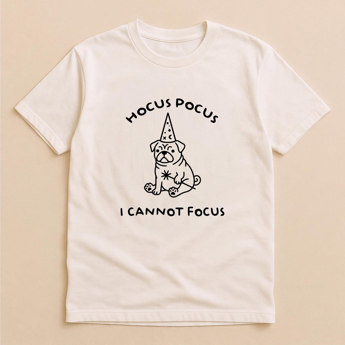 May include: A cream-colored short-sleeve t-shirt featuring a black line-art graphic of a pug wearing a wizard hat with stars and moons. Above the pug is the text HOCUS POCUS and below the pug is the text I CANNOT FOCUS in a simple font.