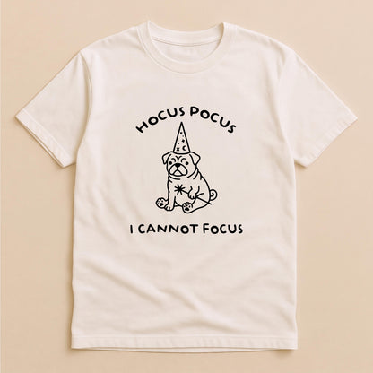 May include: A cream-colored short-sleeve t-shirt featuring a black line-art graphic of a pug wearing a wizard hat with stars and moons. Above the pug is the text HOCUS POCUS and below the pug is the text I CANNOT FOCUS in a simple font.
