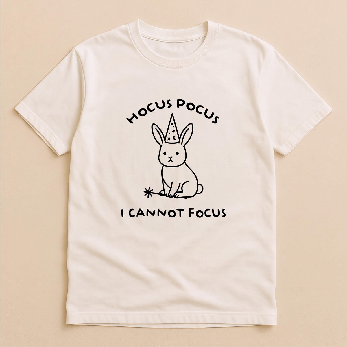 May include: A cream-colored short-sleeve t-shirt featuring a black line-art graphic of a rabbit wearing a wizard hat and holding a magic wand. The text above the rabbit reads HOCUS POCUS and the text below the rabbit reads I CANNOT FOCUS.
