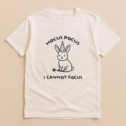 May include: A cream-colored short-sleeve t-shirt featuring a black line-art graphic of a rabbit wearing a wizard hat and holding a magic wand. The text above the rabbit reads HOCUS POCUS and the text below the rabbit reads I CANNOT FOCUS.