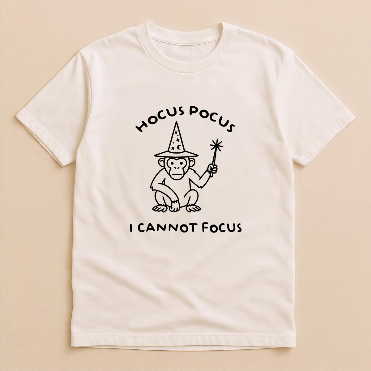 May include: A cream-colored short-sleeve t-shirt featuring a black line drawing of a monkey wearing a wizard hat and holding a magic wand. The text HOCUS POCUS is printed above the monkey, and the text I CANNOT FOCUS is printed below it.