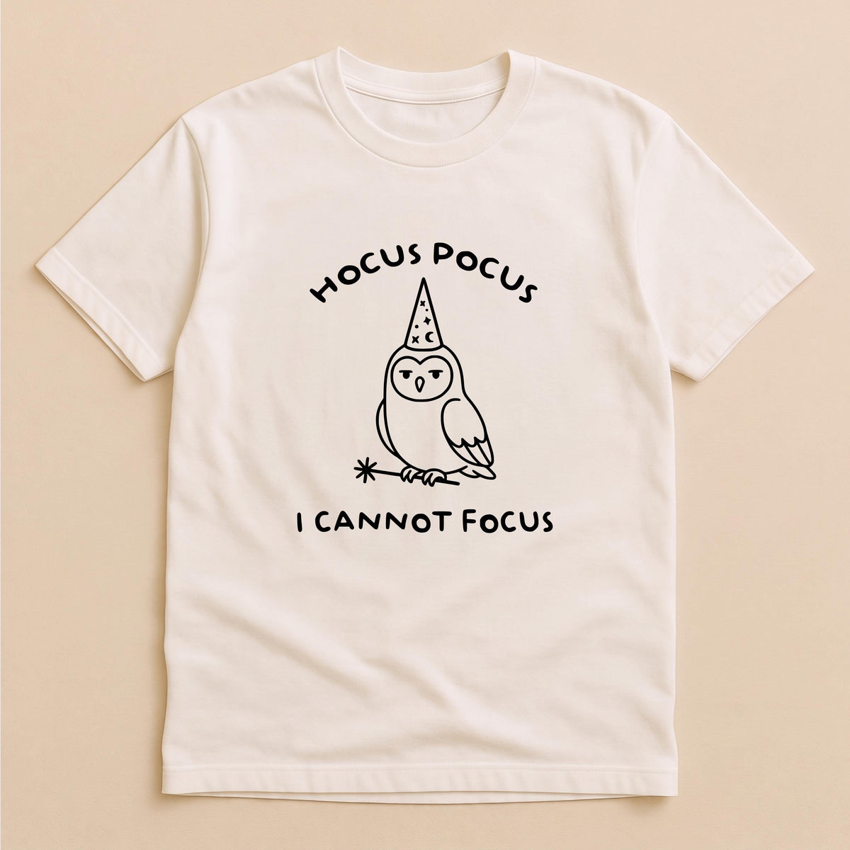 May include: A cream-colored short-sleeve t-shirt featuring a black line-art graphic of an owl wearing a wizard hat and holding a magic wand. The text HOCUS POCUS is printed above the owl, and the text I CANNOT FOCUS is printed below the owl.