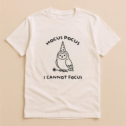 May include: A cream-colored short-sleeve t-shirt featuring a black line-art graphic of an owl wearing a wizard hat and holding a magic wand. The text HOCUS POCUS is printed above the owl, and the text I CANNOT FOCUS is printed below the owl.