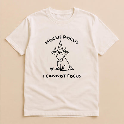 May include: A cream-colored short-sleeve t-shirt featuring a black line drawing of a calf wearing a wizard hat and holding a magic wand. The text HOCUS POCUS is printed above the calf, and the text I CANNOT FOCUS is printed below the calf.