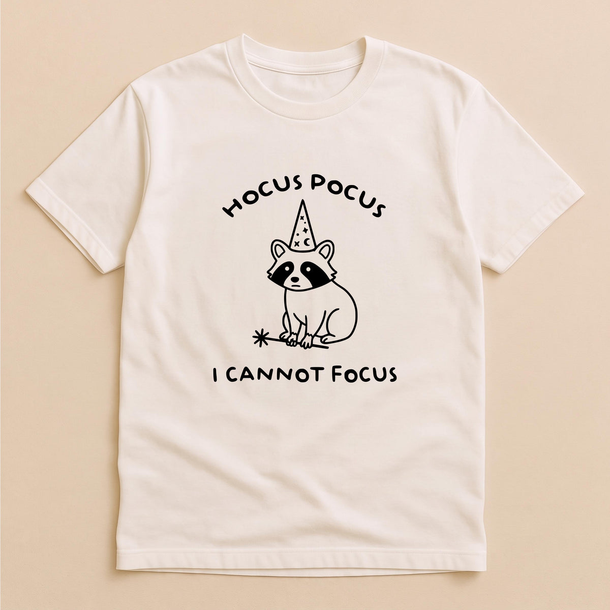 May include: A cream-colored short-sleeve t-shirt featuring a black line-art graphic of a raccoon wearing a wizard hat and holding a magic wand. The text HOCUS POCUS is printed above the raccoon, and I CANNOT FOCUS is printed below the graphic.