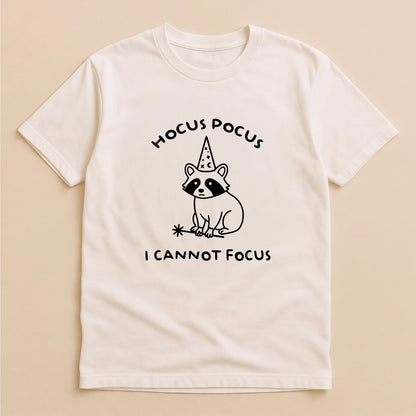 May include: A cream-colored short-sleeve t-shirt featuring a black line-art graphic of a raccoon wearing a wizard hat and holding a magic wand. The text HOCUS POCUS is printed above the raccoon, and I CANNOT FOCUS is printed below the graphic.