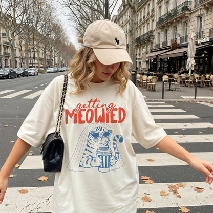 May include: A beige baseball cap with a small dark logo and an oversized cream-colored t-shirt featuring a graphic of a cat wearing sunglasses and a veil, holding a coffee cup, with the red and blue text "getting MEOWIED" printed on the front.