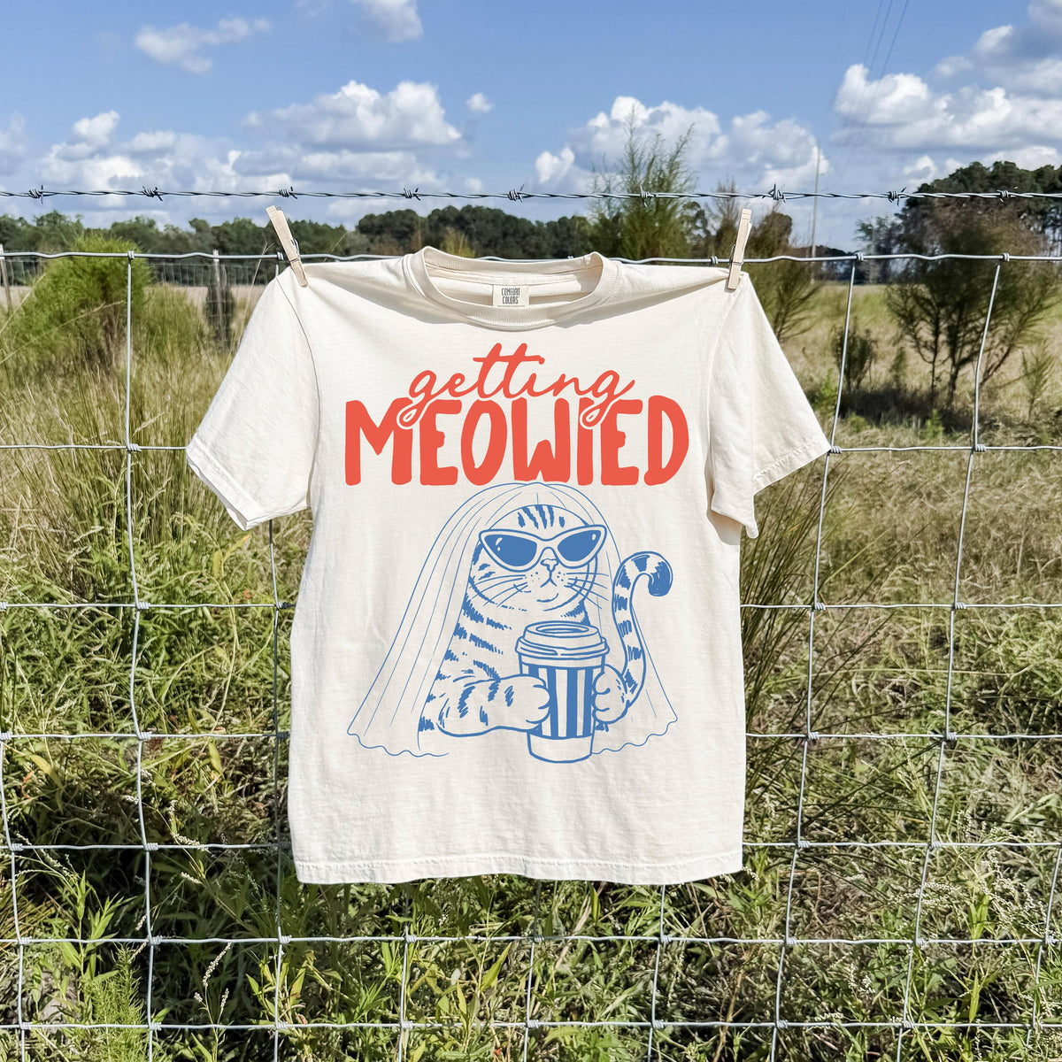 May include: A cream-colored short-sleeve t-shirt featuring a graphic of a cat wearing sunglasses and a bridal veil while holding a coffee cup. The text above the cat reads "getting MEOWIED" in red, with the cat illustration in blue ink.