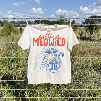May include: A cream-colored short-sleeve t-shirt featuring a graphic of a cat wearing sunglasses and a bridal veil while holding a coffee cup. The text above the cat reads "getting MEOWIED" in red, with the cat illustration in blue ink.