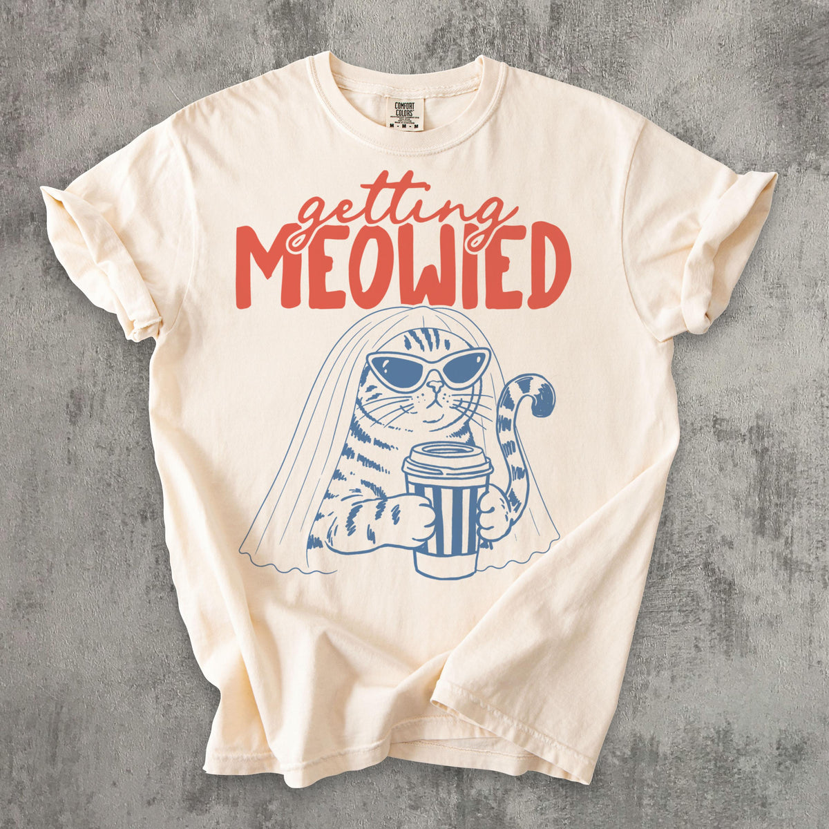 May include: A cream-colored short-sleeve t-shirt featuring a blue line-art graphic of a cat wearing sunglasses and a bridal veil while holding a coffee cup. Above the cat, the text reads "getting MEOWIED" in a bold, red, stylized font.