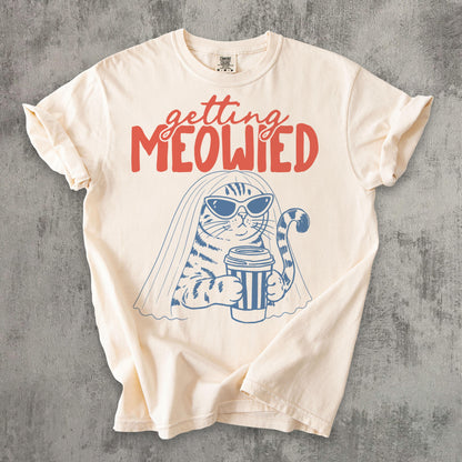 May include: A cream-colored short-sleeve t-shirt featuring a blue line-art graphic of a cat wearing sunglasses and a bridal veil while holding a coffee cup. Above the cat, the text reads "getting MEOWIED" in a bold, red, stylized font.