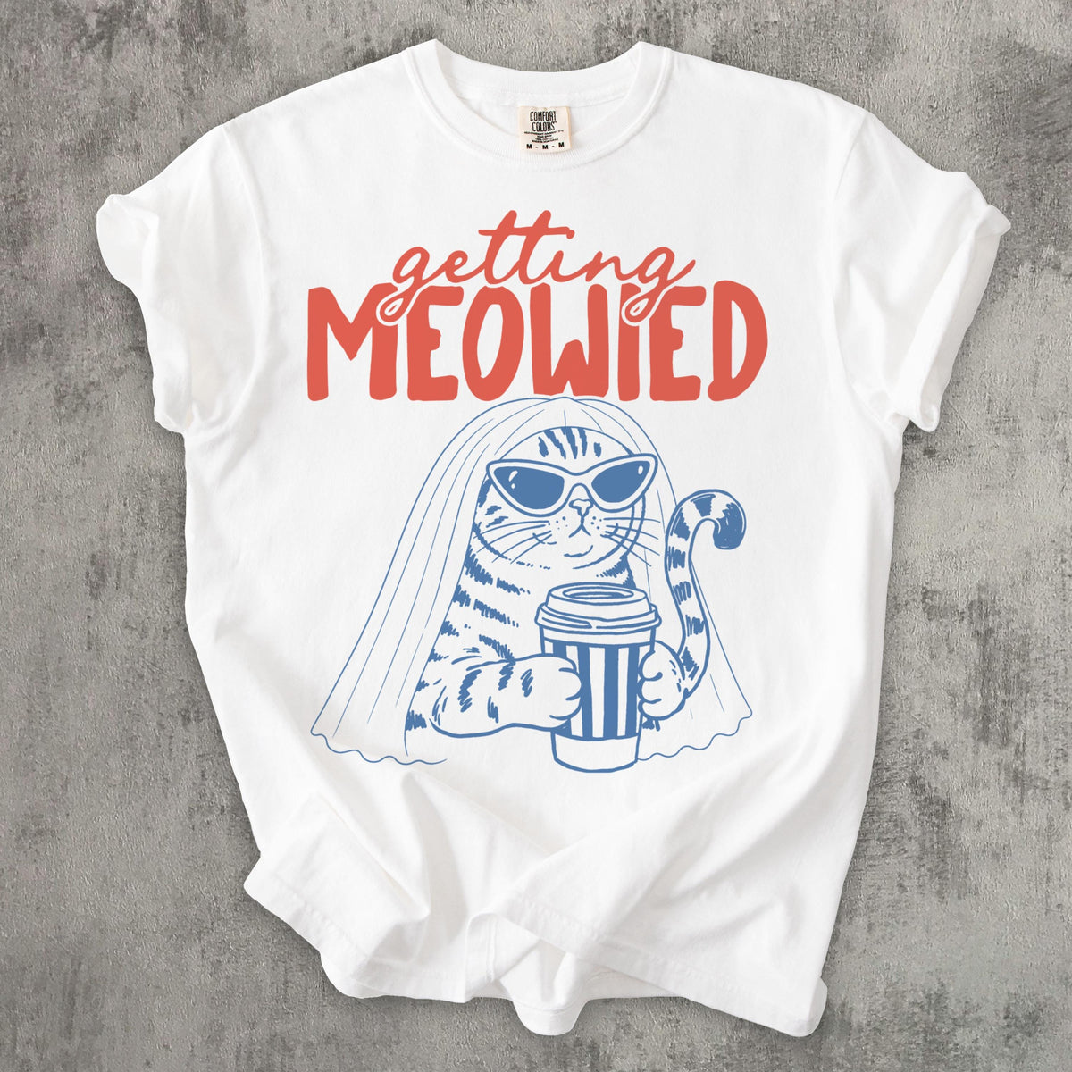 May include: A white t-shirt featuring a blue line-art illustration of a cat wearing sunglasses and a bridal veil while holding a striped coffee cup. Above the cat, the text "getting MEOWIED" is printed in a bold, coral-red script and block font.