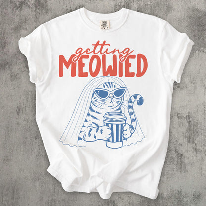 May include: A white t-shirt featuring a blue line-art illustration of a cat wearing sunglasses and a bridal veil while holding a striped coffee cup. Above the cat, the text "getting MEOWIED" is printed in a bold, coral-red script and block font.