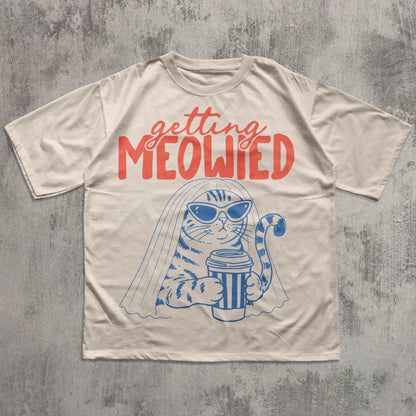 May include: A beige short-sleeve t-shirt featuring a blue graphic of a cat wearing sunglasses and a veil while holding a coffee cup. Above the cat, the text "getting MEOWIED" is printed in a bold, red, stylized font across the chest area.
