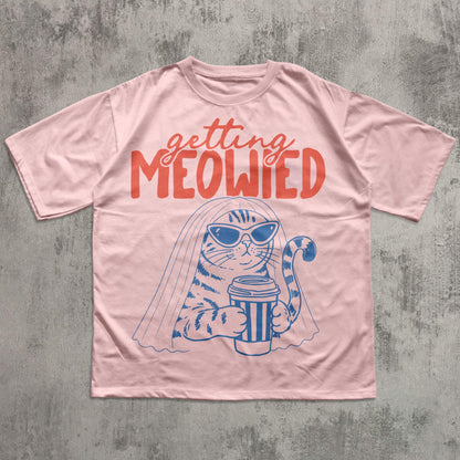 May include: A light pink short-sleeve t-shirt featuring a blue graphic of a cat wearing sunglasses and a veil while holding a coffee cup. Above the cat, the text reads "getting MEOWIED" in a bold, orange, stylized font across the chest.
