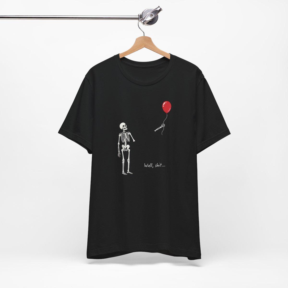 May include: A black short-sleeve t-shirt featuring a white graphic of a skeleton looking at a red balloon floating away with a microphone attached to the string. The text "Well, shit..." is printed in white below the skeleton on the front of the shirt.