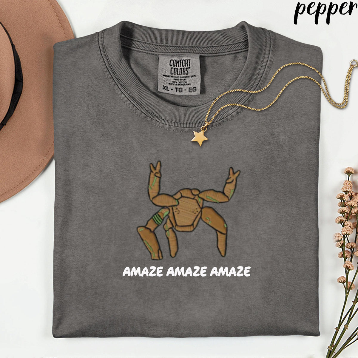 May include: A charcoal gray t-shirt with a Comfort Colors label at the neck. The shirt features a graphic of a brown, multi-legged creature with green accents and the text "AMAZE AMAZE AMAZE" printed in white below the illustration.
